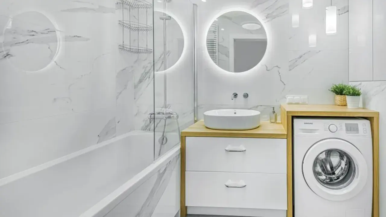 Nowoczesne, małe łazienki z wanną, umywalką nablatową i pralką. Marmurowe płytki i okrągłe lustro z podświetleniem tworzą elegancki styl.