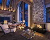 Przytulny salon w stylu chalet z kamiennym kominkiem, drewnianymi belkami i dużymi oknami.