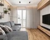 Przytulny salon w bloku z szarą kanapą, drewnianą podłogą i nowoczesnym telewizorem. Na ścianie wiszą czarne półki z książkami i roślinami.