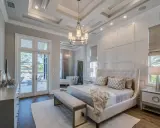 Elegancki master bedroom z łóżkiem z wysokim zagłówkiem, ozdobnymi poduszkami i narzutą. Widok na taras z basenem.