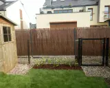 Nowe maliny w ogrodzie aranżacje. Krzewy rosną na rabacie, otoczone korą i kamieniami. Obok drewniana szopa i furtka.