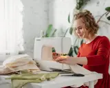 Kobieta w czerwonej sukience pracuje przy maszynie do szycia, dbając o naprężenie nici. Na stole leżą kolorowe tkaniny i nożyczki.