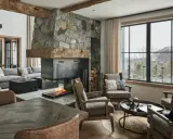 Przytulny salon w stylu modern farmhouse z kamiennym kominkiem, drewnianymi belkami i widokiem na ośnieżone góry.