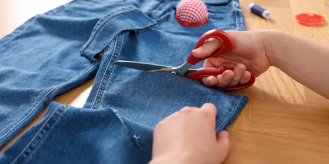 Ręce przycinają za długie spodnie dżinsowe, aby je skrócić. Na stole leżą też nici i poduszeczka na szpilki.