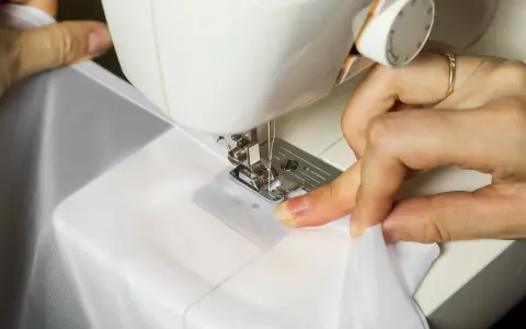 Ręce prowadzą białą tkaninę pod stopkę maszyny do szycia, pokazując, jak obszyć brzegi firanki.