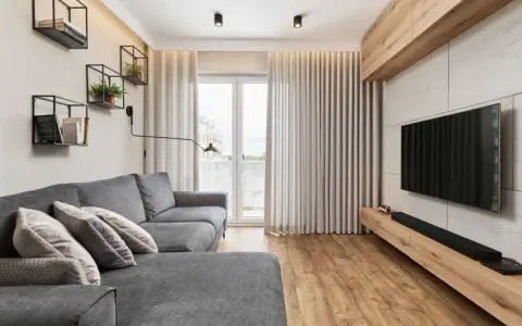 Przytulny salon w bloku z szarą kanapą, drewnianą podłogą i nowoczesnym telewizorem. Na ścianie wiszą czarne półki z książkami i roślinami.