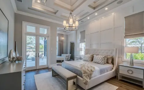 Elegancki master bedroom z łóżkiem z wysokim zagłówkiem, ozdobnymi poduszkami i narzutą. Widok na taras z basenem.