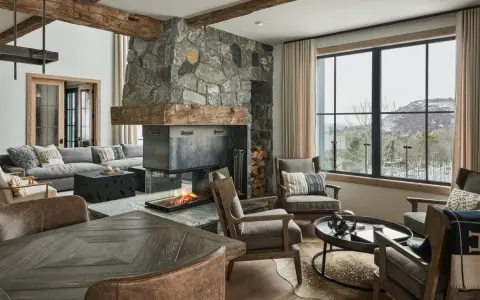 Przytulny salon w stylu modern farmhouse z kamiennym kominkiem, drewnianymi belkami i widokiem na ośnieżone góry.