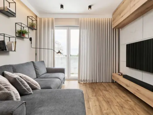 Przytulny salon w bloku z szarą kanapą, drewnianą podłogą i nowoczesnym telewizorem. Na ścianie wiszą czarne półki z książkami i roślinami.