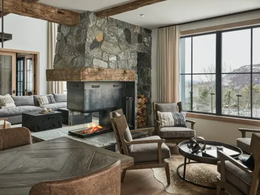 Przytulny salon w stylu modern farmhouse z kamiennym kominkiem, drewnianymi belkami i widokiem na ośnieżone góry.