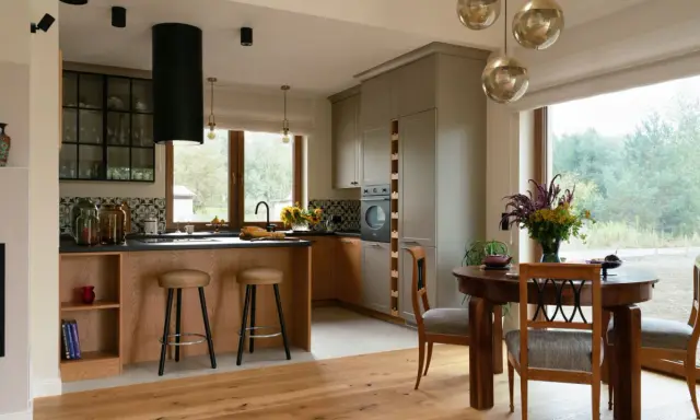 Przytulna kuchnia w stylu modern farmhouse z drewnianą wyspą, szarymi szafkami i dużym oknem z widokiem na zieleń.