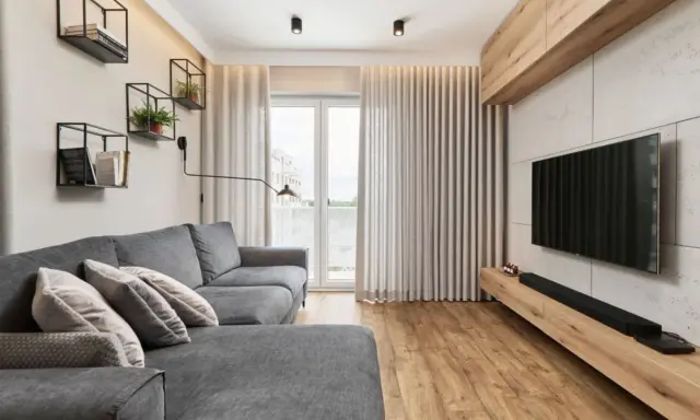 Przytulny salon w bloku z szarą kanapą, drewnianą podłogą i nowoczesnym telewizorem. Na ścianie wiszą czarne półki z książkami i roślinami.