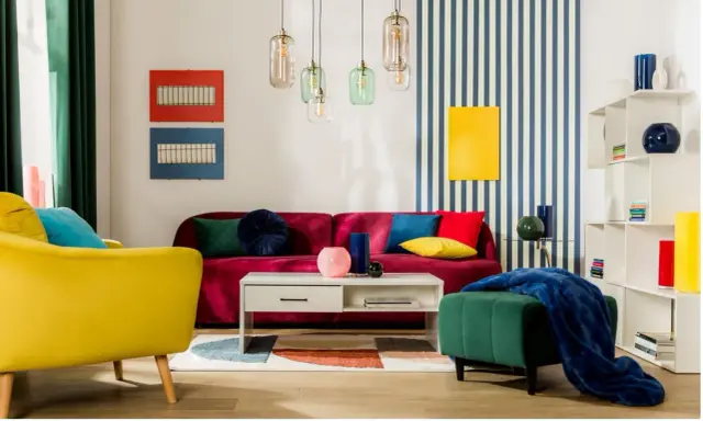 Salon w stylu memphis z kanapą w kolorze fuksji, żółtym fotelem i zieloną pufą. Kolorowe poduszki i geometryczne dekoracje.
