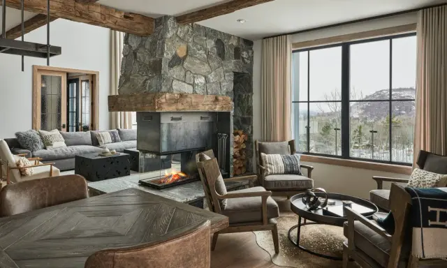 Przytulny salon w stylu modern farmhouse z kamiennym kominkiem, drewnianymi belkami i widokiem na ośnieżone góry.