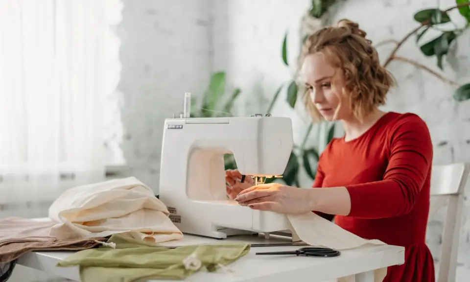 Kobieta w czerwonej sukience pracuje przy maszynie do szycia, dbając o naprężenie nici. Na stole leżą kolorowe tkaniny i nożyczki.