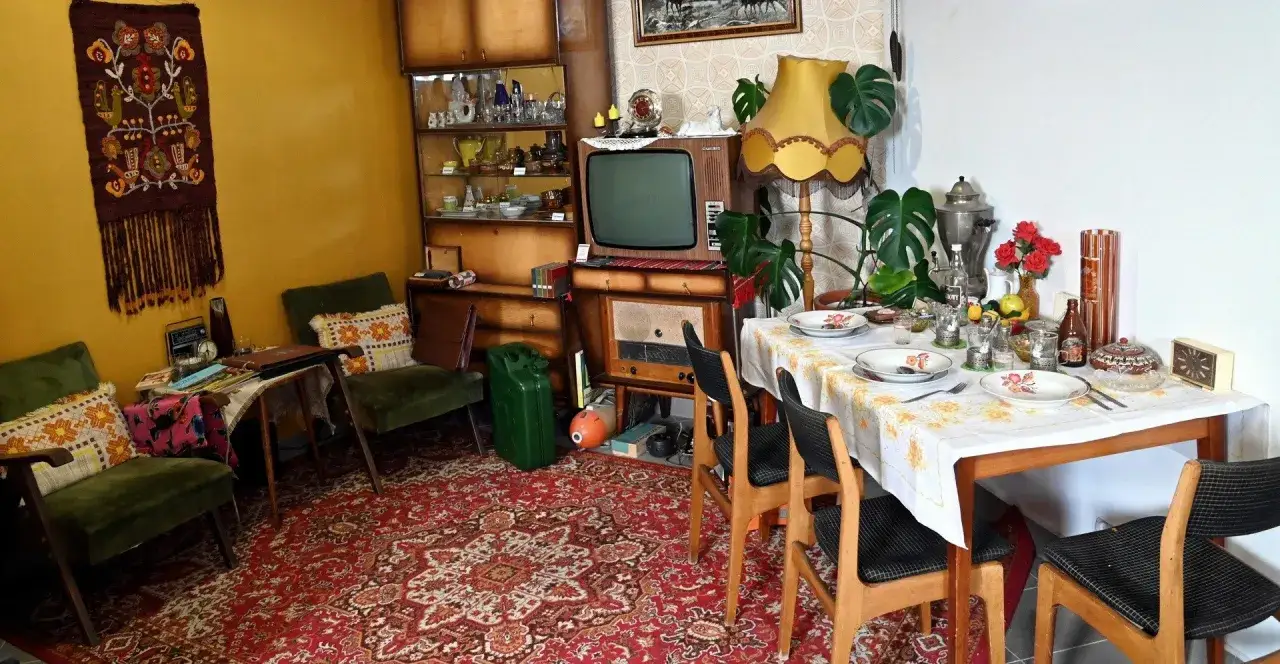 Wnętrze mieszkania PRL: zielone fotele, dywan, meblościanka, telewizor, radio, st&oacute;ł z zastawą i kwiatami.