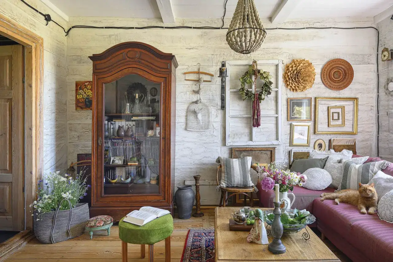 Przytulny salon w stylu cottage core. Drewniana witryna, kanapa z kotem, bukiet kwiat&oacute;w i mn&oacute;stwo bibelot&oacute;w tworzą sielski klimat.